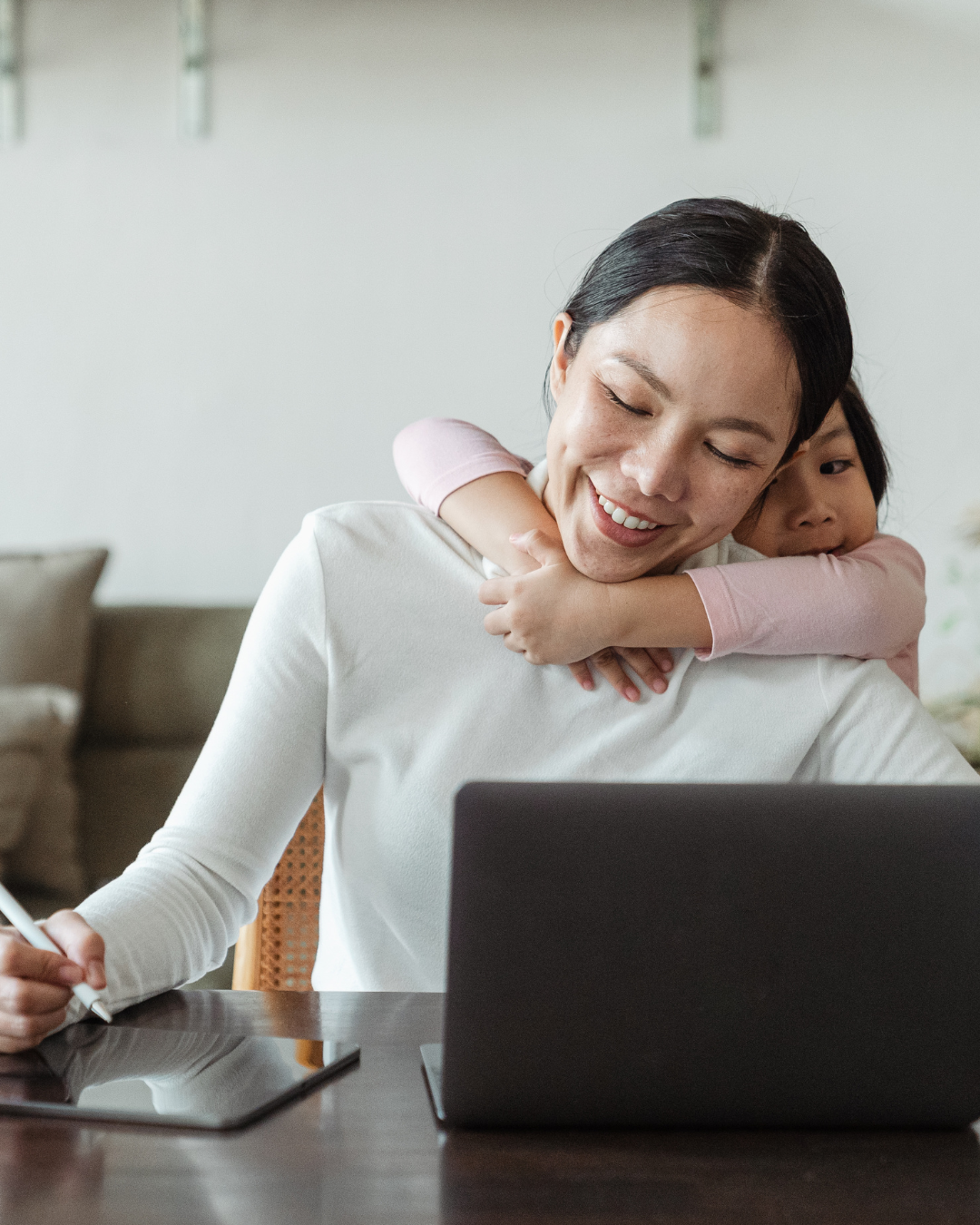 A woman at her desk trying to work while her daughter hugs her back, then they both smile.