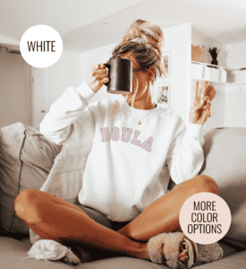 White woman with slippers and a messy bun holding a coffee mug over her face with a peace sign up wearing an oversized white sweatshirt that says doula across the chest in pink varsity font.